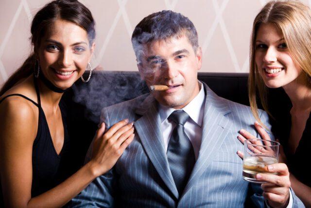Portrait of successful man smoking a cigar holding whisky with pretty women near by